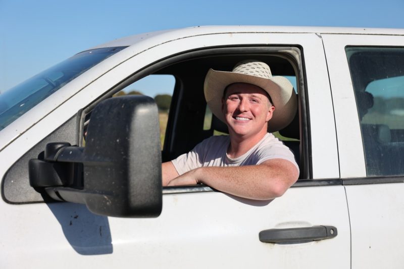 A young man in a cowboy hat smiles from behind the wheel of a white pickup truck.