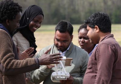 A group of students and their professor look at control screen of drone in preparation for flight.