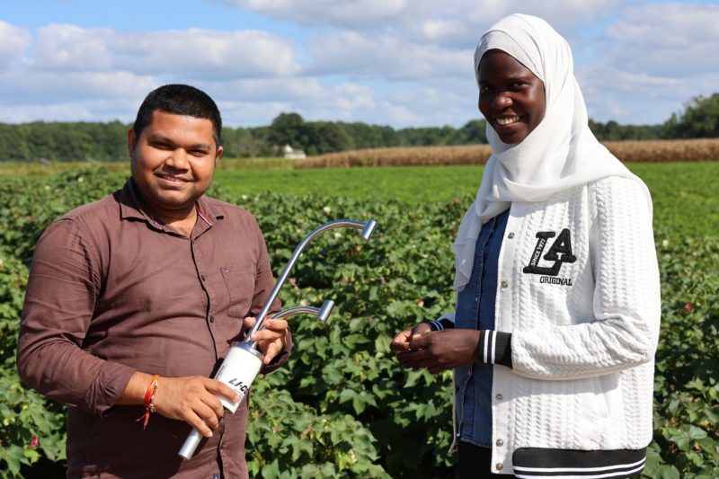 Man in brown shirt holds water sensor in field with young woman in head scarf standing next to him.