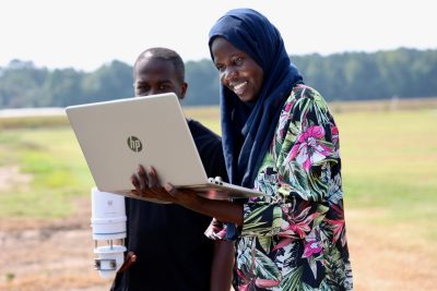 Young woman with head scarf holds laptop in field with young man carrying a weather station