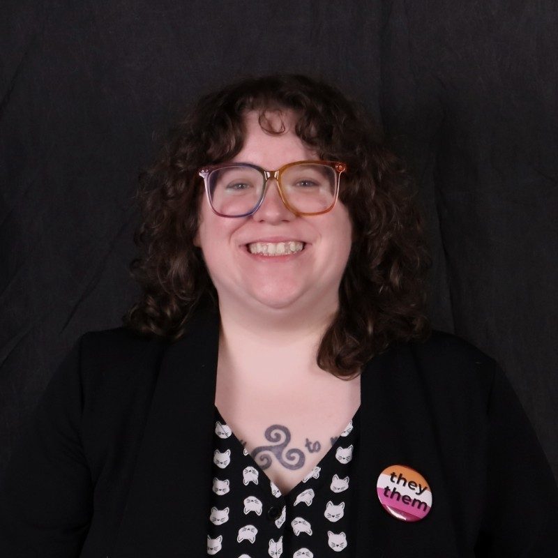 Alexandra Pirkle director of communications and marketing for Office of Inclusion and Diversity is seen standing against a dark grey background dark curly hair, eye glasses and smiling. They have a black overcoat on with a black shirt that features a pattern of cat faces.