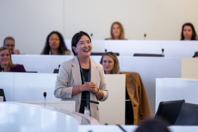 an asian woman in a dark shirt and a beige sweater smiles at something off camera