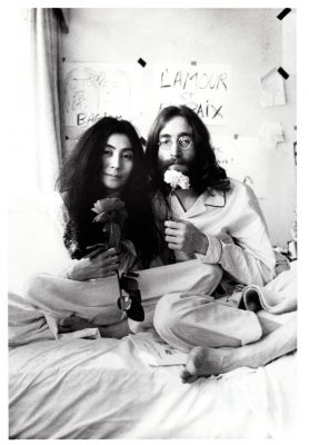 Yoko Ono and John Lennon sit in a bed holding flowers