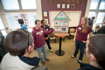 Three Virginia Tech students stand on either side of a small model house while other students look on.