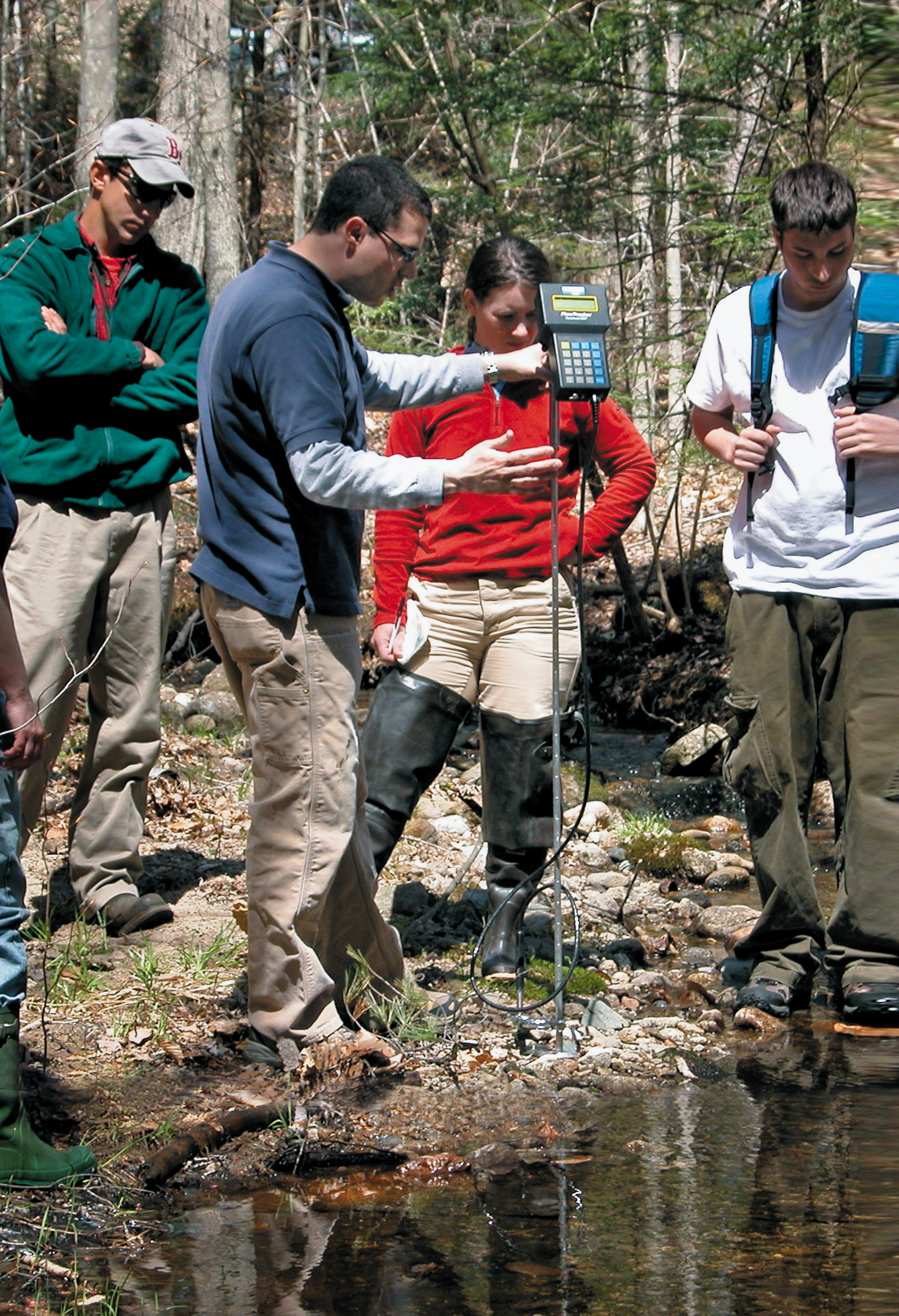 Four people standing next to a forest stream. One holds a water meter.