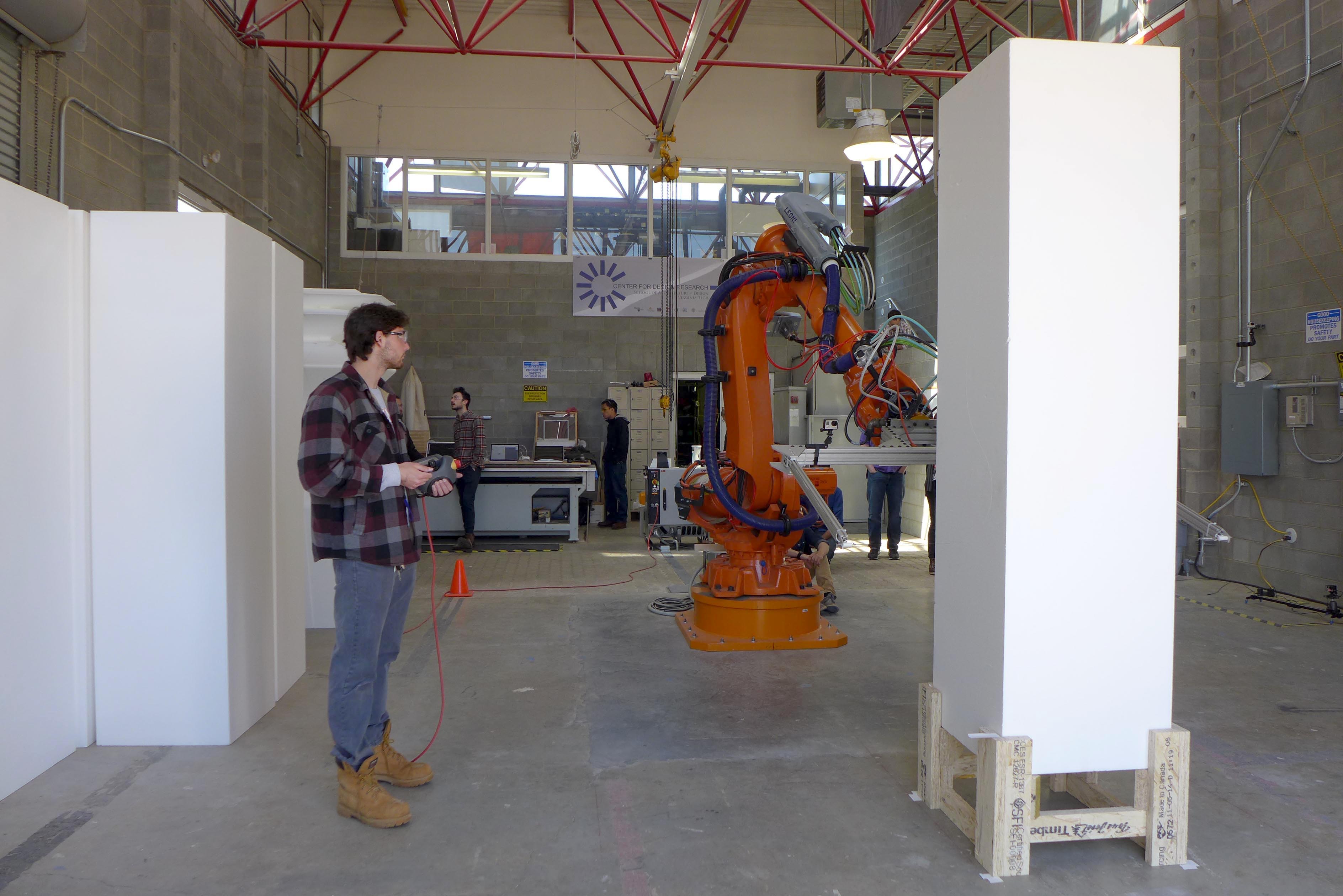 A young man controls a large industrial robot with a remote control as it prepares to cut a 7-foot styrofoam block.