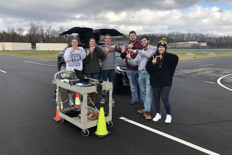 Six engineering graduate students stand in front of a car and use their hands to make the "VT" shape.