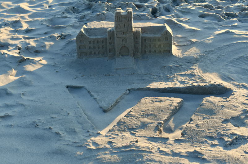 A sandcastle in the shape of Burruss Hall, sits behind the VT athletic mark, written in sand.