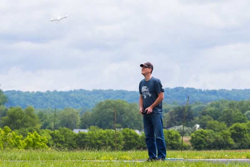 A man stands in a field holding a controller while a small white drone flies nearby