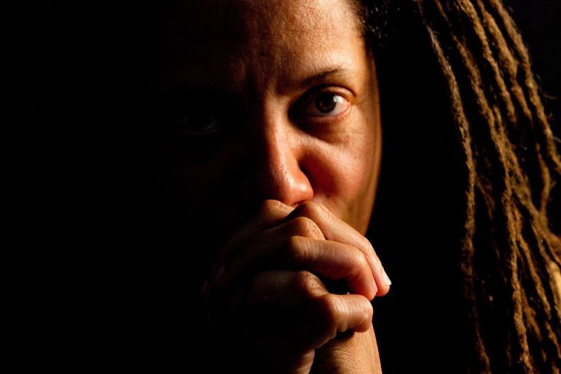 A close up portrait of Nikky Finney with her hands folded in front of her face, looking into the camera.