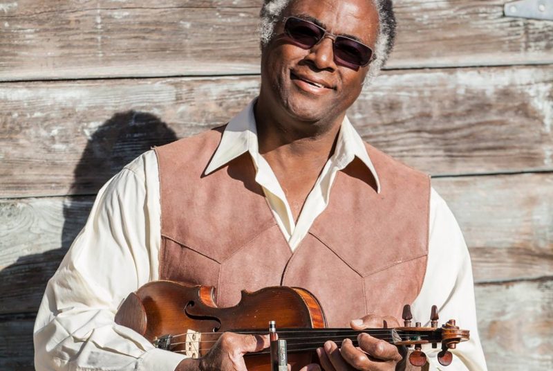 Musician Earl White wears sunglasses and a tan vest over a white shirt while posing outdoors in front of a wooden wall holding his fiddle.