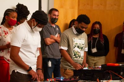 Two students play with the turntables, as the entire class is standing behind them to listen.