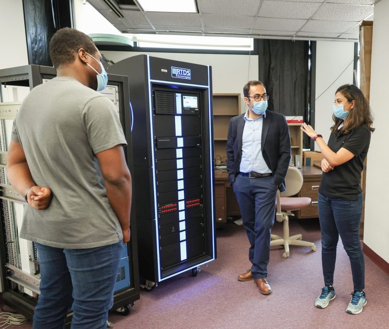 Three graduate students gather around a tall black box with a lighted display reading "RTDS technologies."