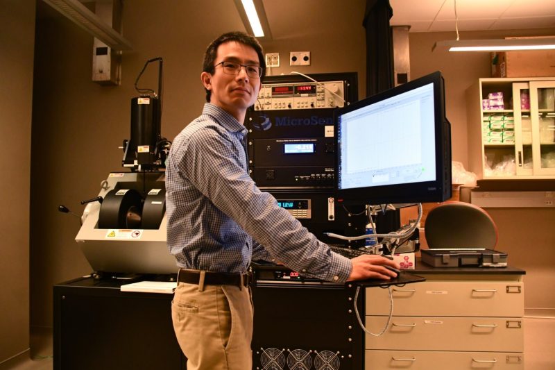 Satoru Emori stands at a sensor machine and computer in one of his labs at the ICTAS 2 building. He's wearing slacks and a checkered shirt. Photo for Virginia Tech by Steven Mackay. 