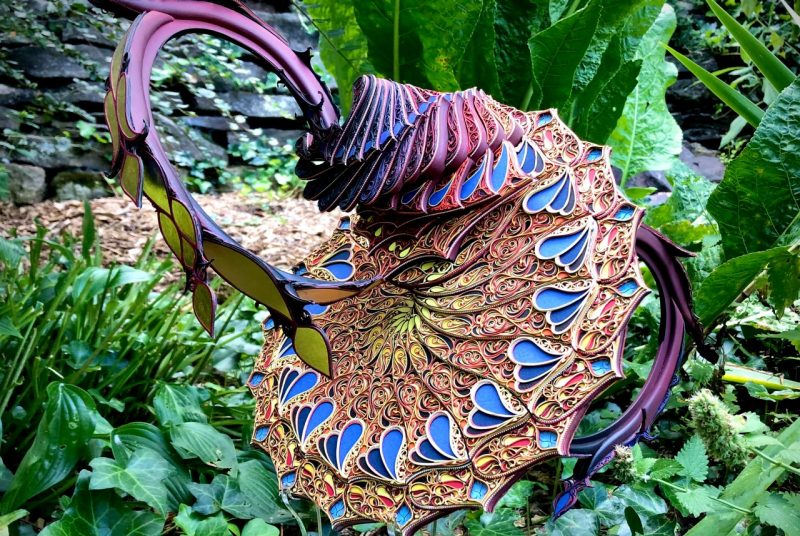 Similarity of Being is an artwork made from cut paper, wood, gold leaf, botanicals, water, and PVC. It looks like an exotic flower - blue and purple petal shapes fan around a circular base, while a piston-looking structure rises from the top.