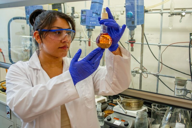Female student holds and looks at a glass beaker filled with sediment and liquid.