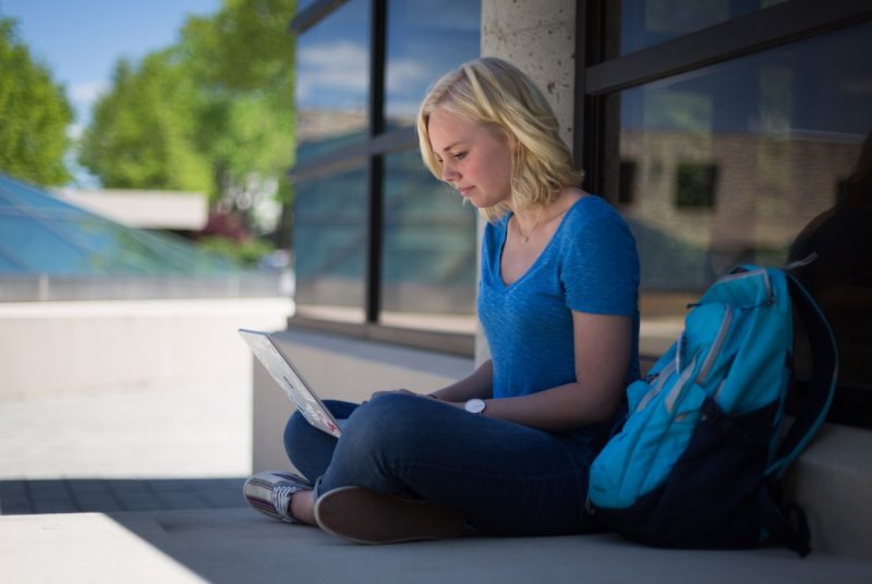 Student sits on the ground outside, with her back against the front windows of a building and looking at a laptop which is open in her lap.