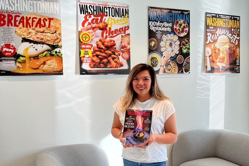 Jade Womac stands and holds a copy of the Washingtonian magazine. Behind her are four large prints of Washingtonian covers that hang on a wall.