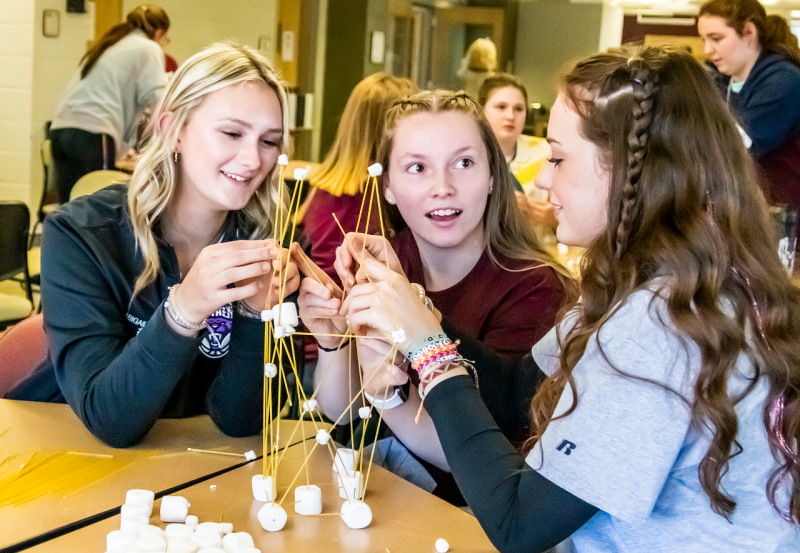 Three high school girls build a building frame out of spaghetti noodles and marshmallows.
