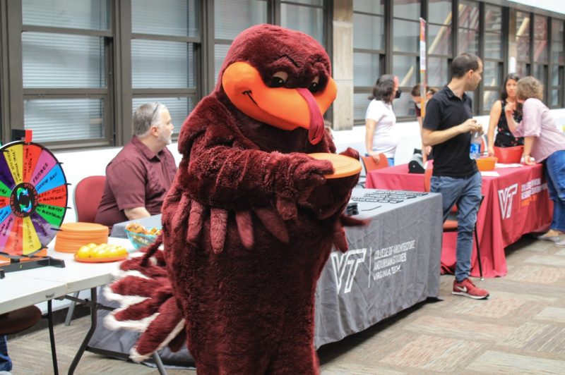 The Hokie Bird gets ready to toss a Frisbee, while in the background people sit at booths at the First Year Finish event