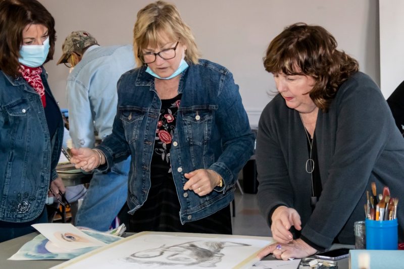 Three women stand looking at a drawing laying on a table in front of them.