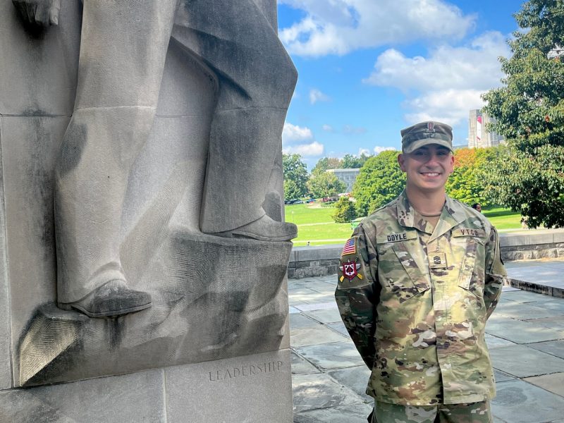 Cadet Doyle is smiling next to the lower half of the Leadership pylon monument. Campus and Burruss Hall are in the background with white clouds and blue sky. The leaves are starting to change.
