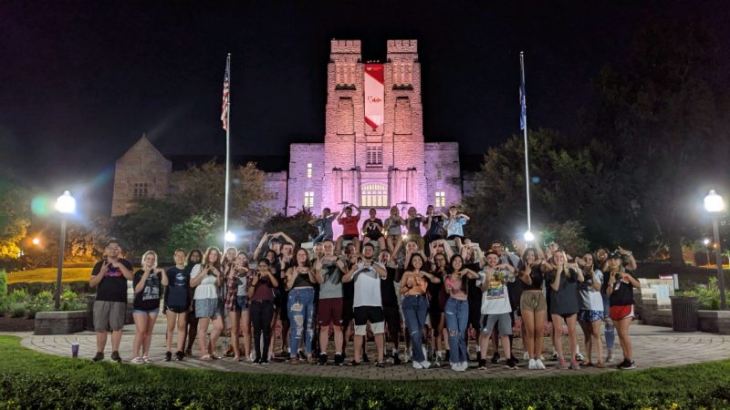 At night, in front of a brightly lit Burruss Hall, a large group of transfer students make the VT sign with their hands.