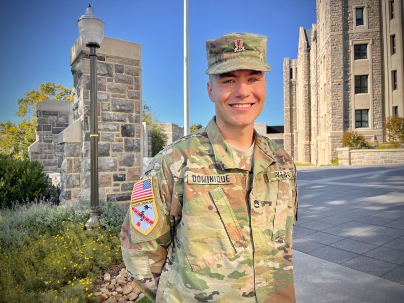  Cadet Dominique smiles and stands on Upper Quad with Pearson Hall West and the flagpole in the background.