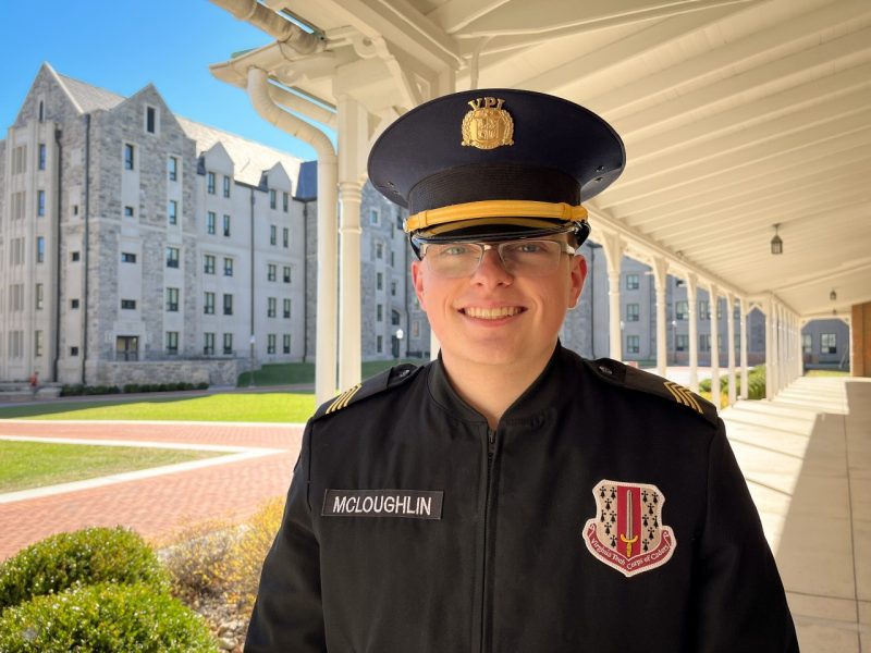 Cadet McLoughlin stands in uniform smiling on Upper Quad with Pearson Hall West and the porch of Lane Hall in the background.