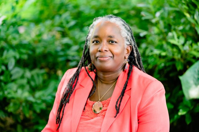Vice President for Strategic Affairs and Diversity, Dr. Menah Pratt photographed in the Hahn Garden in Summer of 2022. The photo is a close frame. She is wearing a coral-color suit. The background is composed of plants, green out-of-focus. 