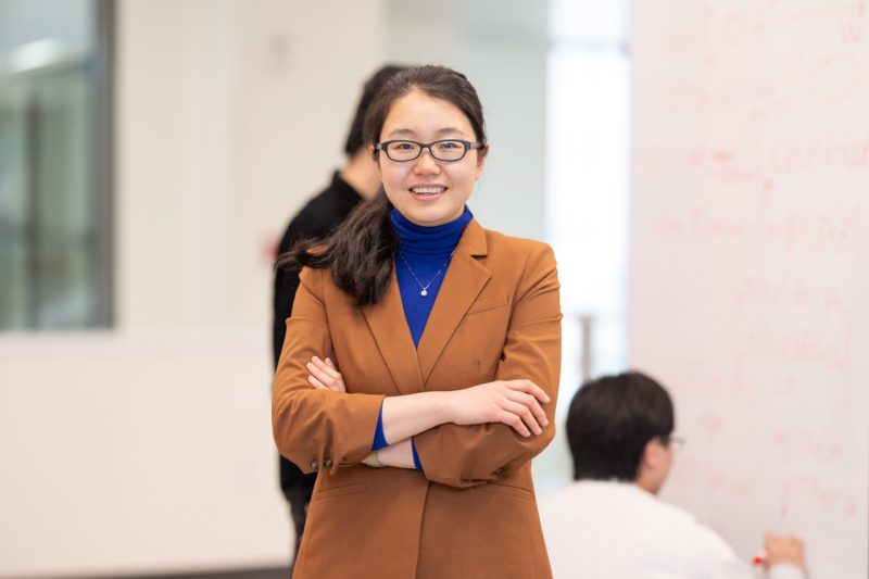 Ruoxi Jia stands with arms crossed and smiling at the camera while students work in the background
