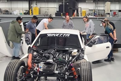 Students stand around a white sports car with an exposed engine on a factory floor. 