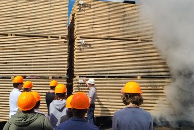 A group of students stands near a large pallet of wood with steam rising from the right side.