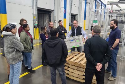 A man holds a plastic box as others look on, while standing on a factory floor.