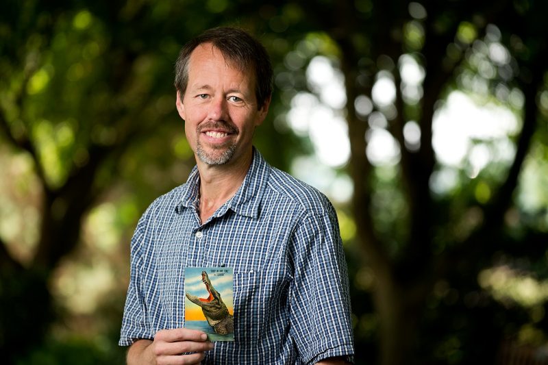 Mark Barrows wears a blue plaid shirt and holds a vintage postcard of an alligator.