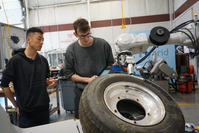 Two men stand side by side looking at a small digital tablet, while in the foreground a large vehicle tire is held by a laboratory apparatus. 