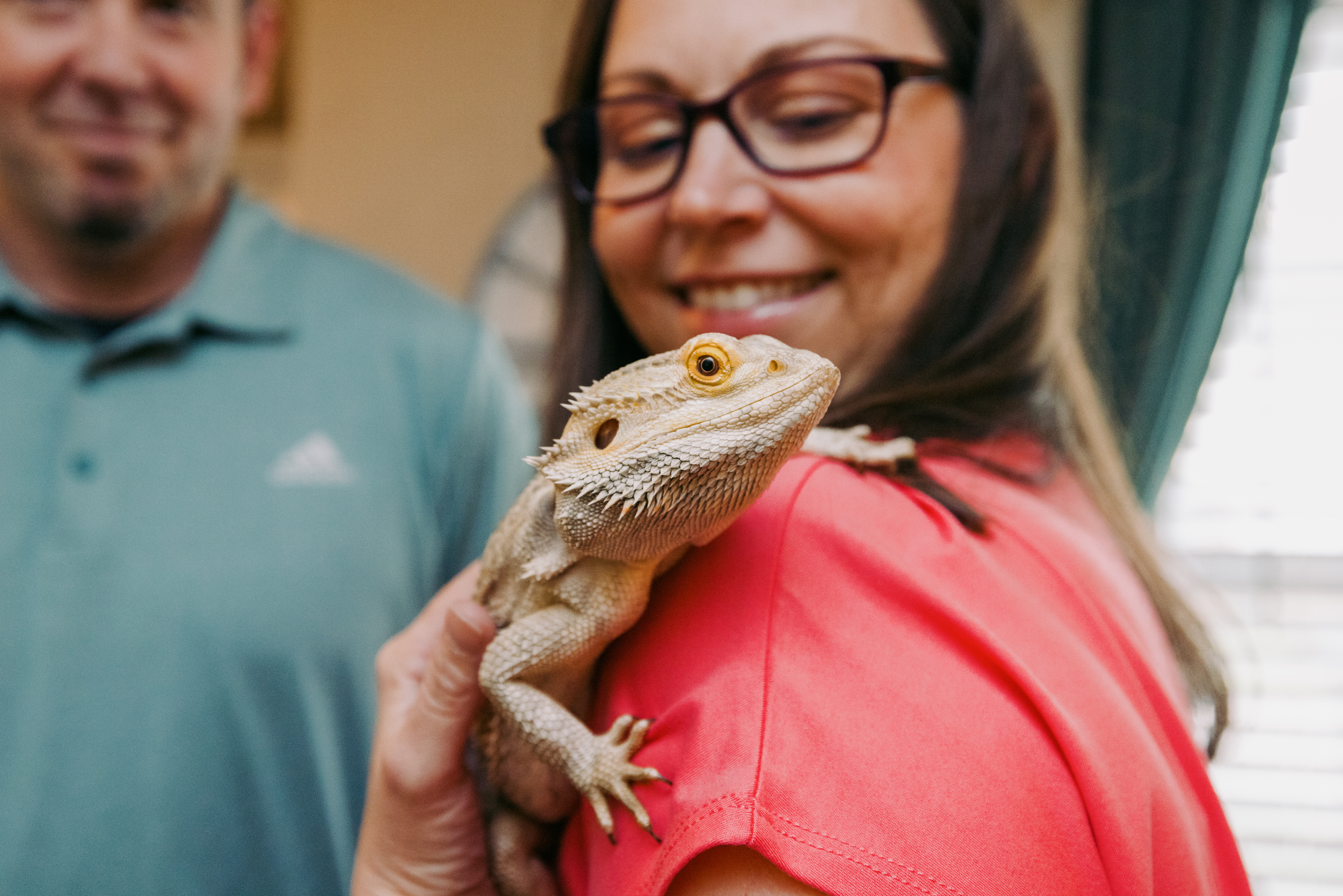 Holly Williams smiles as a pale yellow bearded dragon named Drax sits on her shoulder; her husband Daron looks on.