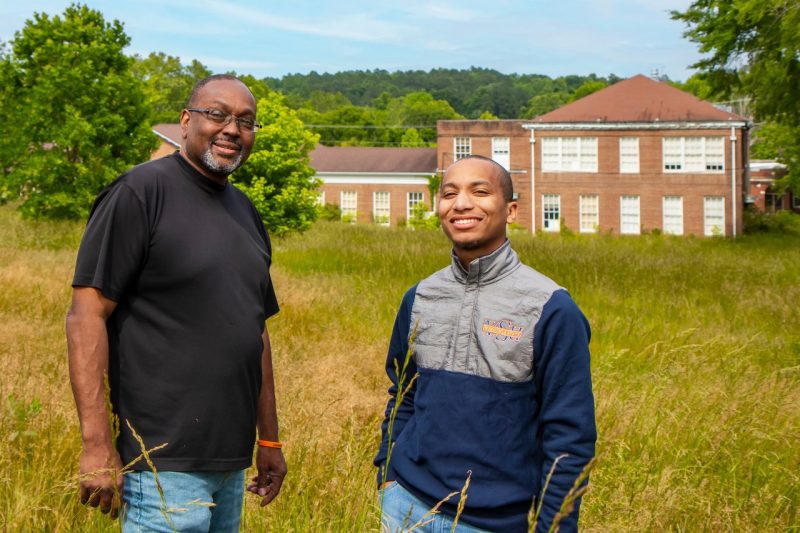 Two men stand in a field of high grass with an old brick school building in the background.