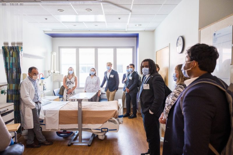 A group of students stand in a semi-circle inside a hospital room as a scientist from the NIH talks with them about various features of the room.