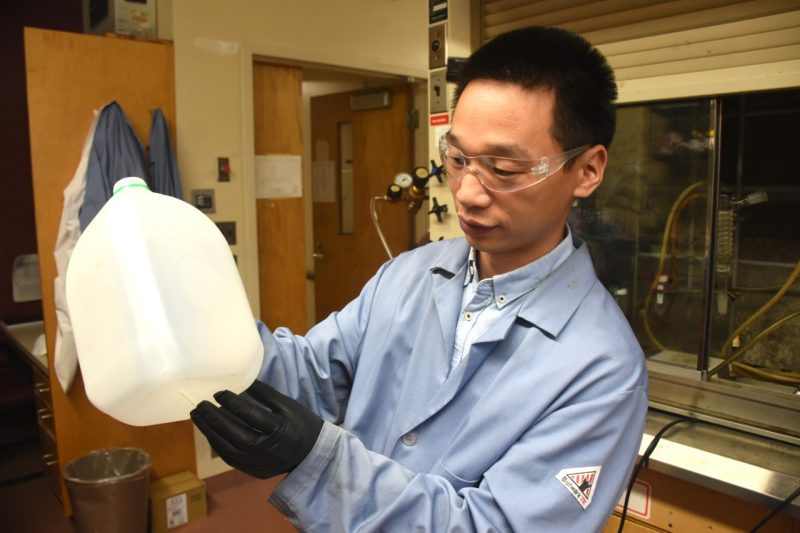 A man with dark hair wears a lab coat and holds a water jug.