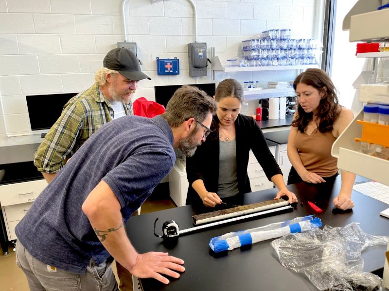 Inside a geosciences lab, two men and two women examine a long half-pipe holding samples core sediment collected in Oregon.