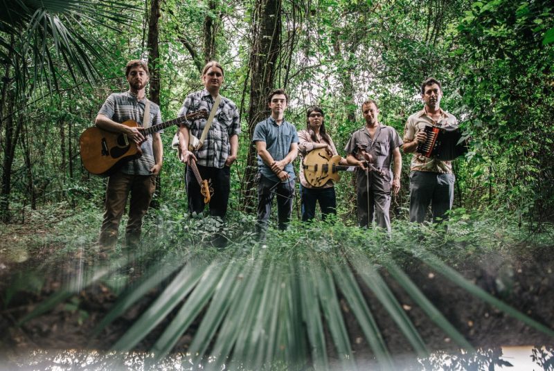 The six musicians with the group Lost Bayou Ramblers stand in the middle of a lush, green forest, surrounded by trees and foliage, while holding their instruments.