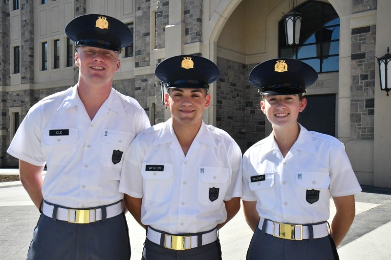 Cadets in white uniform shirts stand smiling in front of the Corps Leadership and Military Science Building.