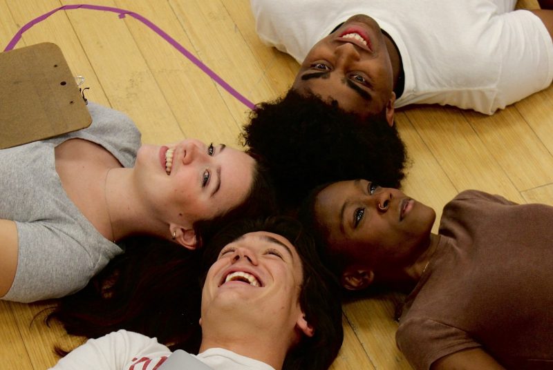 Four students lie on the floor with their heads touching each other.