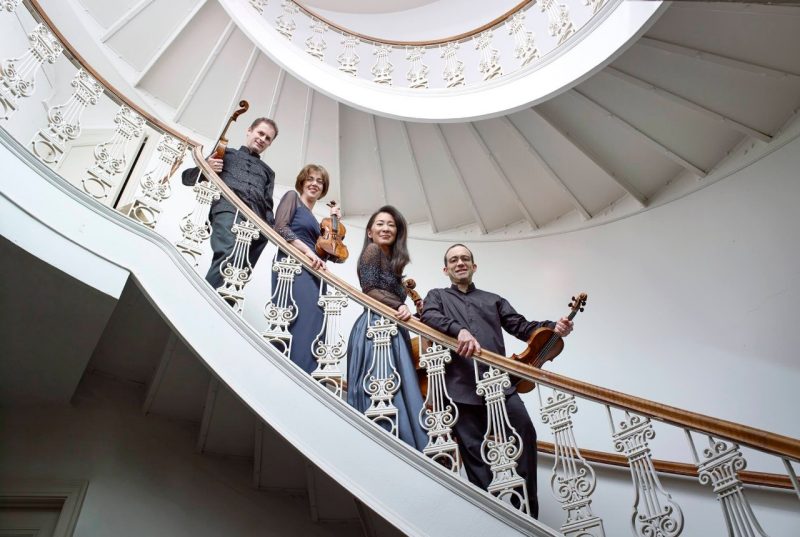 The members of the classical music ensemble Brentano String Quartet stand on the steps of a very ornate staircase. The members - two men and two women - each hold their instruments and each stands on their own step.