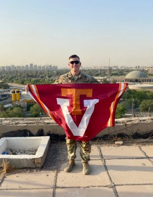 Kase smiles while holding a Virginia Tech flag. He is wearing his camouflage uniform and sunglasses in a desert setting with a Middle East city in the background.