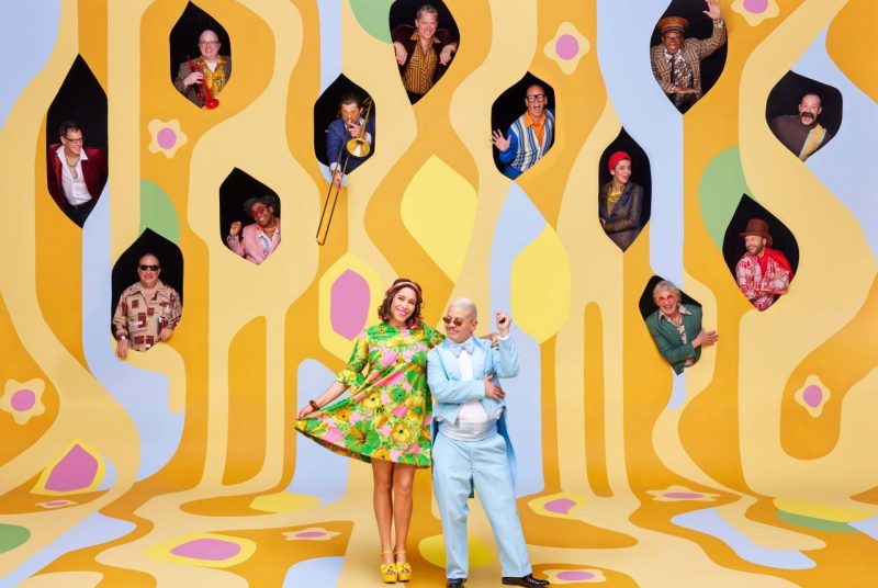 The members of Pink Martini wear '60s-inspired outfits and dance in front of a multicolored '60s-inspired backdrop, some of them holding their instruments.