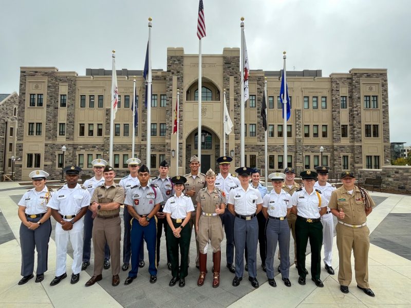 Student leaders in a mix of cadet uniforms from various schools stand smiling in front of the building.
