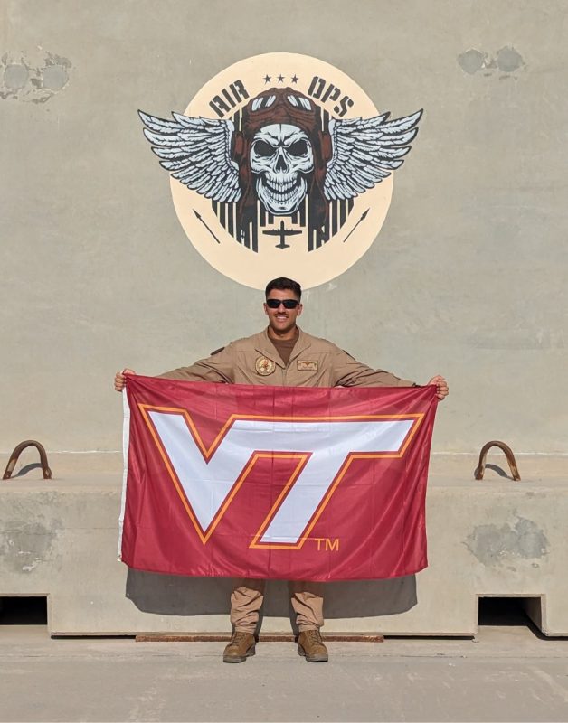 U.S. Navy Lt. Gupta smiles while holding a Virginia Tech flag. He is wearing his beige flight suit and sunglasses and standing below painted artwork that reads “Air Ops.”