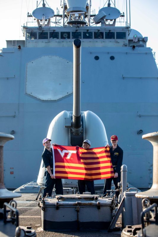 Three servicemembers in blue coveralls stand holding a striped VT flag in front of the ship’s bridge on the deck of a U.S. Navy cruiser. 